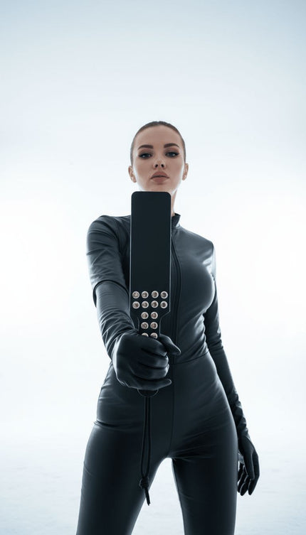 Жена в латексов костюм държи кожен bdsm падъл с метални капси